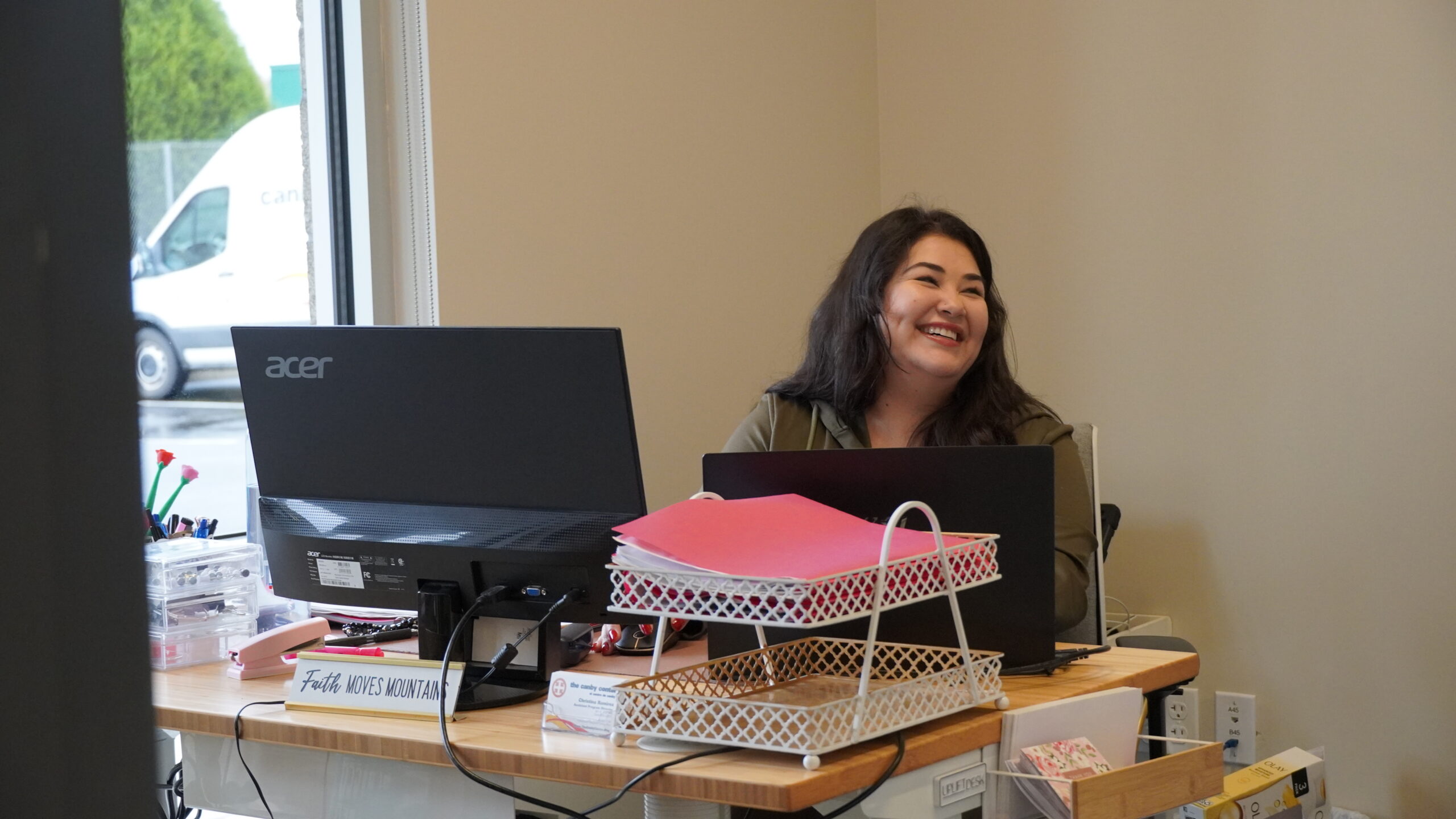 Woman sitting at her desk, smiling behind a computer and folders. Employment at To:gether Canby.